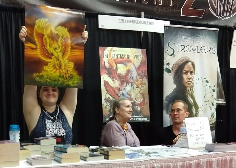 Mercedes Lackey (center) at the Zombie Orpheus Entertainment stall. The person at left holds up a print of one of Larry Dixon's paintings.